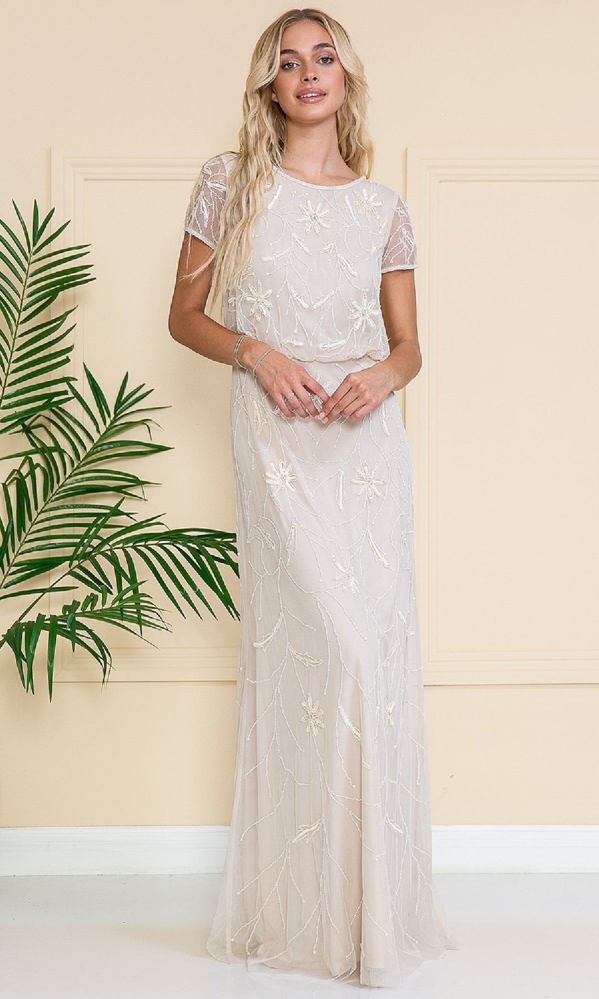 A woman in the Blouson Long Formal Dress: Amelia Couture IN004, featuring delicate floral embellishments, stands indoors beside a green leafy plant and a cream-colored wall.